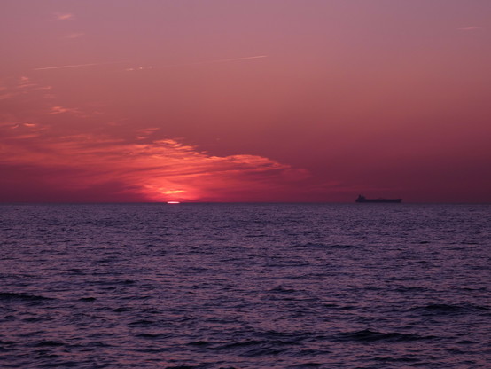 Sehr roter Himmel, die Sonne kaum noch zu sehen, kurz vor dem völligen Versinken "in der See" (d.h. hinter den Horizont in der Ostsee), westlich. Das gleiche Schiff, wie im Bild davor, ist rechts am Horizont zu sehen. Die See sieht metallisch blau aus. Nur einzelne Wolkenfetzen am Himmel, die von der Sonne angestrahlt werden.