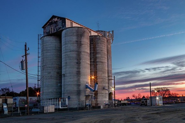Dixon Grain Elevator

This is an image of a Grain Elevator located in Dixon California.
Grain elevators to me are a lot like boats. They seem to have their own style and character. This one has some kind of warehouse or office located on top of the large cylindrical towers. 
There are other buildings in the background and the foreground is an empty parking lot. 
The sun is just starting to breach the horizon off to the right of the image creating a nice warm pinkish glow.