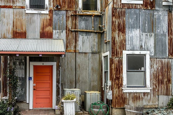 Orange Door

This is an image that I shot in the process of trying to emulate a couple of classic photographers, William Quisenberry and Walker Evans. Both are known to shoot images that are pretty much two dimensional without much depth.
This is the back of an old building found out in the Sacramento River Delta in the town of Locke. 
The siding on the building is all corrugated steel, some of the pieces are showing different levels of rust giving it a very industrial look. 
There are some odds and ends on the ground like a hose and an old planter along with various other items.
What caught my eye on this image is the reddish/orange door in the lower left hand corner.