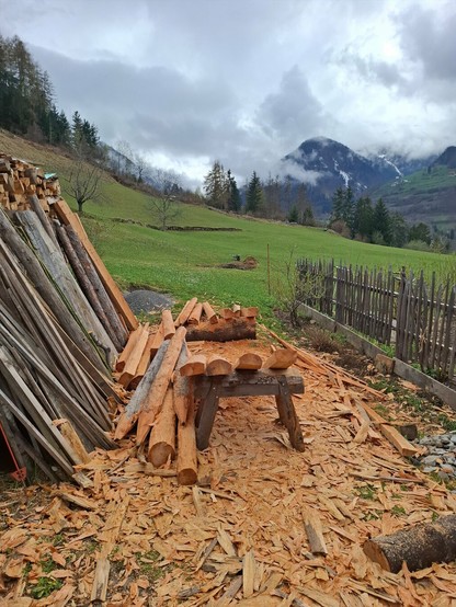 A rural outdoor woodworking area set against a mountainous backdrop. In the foreground, freshly stripped logs—mostly larchwooden fence posts with their sapwood copped—are scattered around a sturdy wooden sawhorse. Wood shavings and bark chips densely cover the ground. On the left, a neat stack of similarly prepared timber leans against a structure. A rustic wooden fence runs along the right side, marking the edge of a grassy field. In the background, gently sloping meadows lead to forested hills and cloud-draped mountains. The overcast sky casts a soft, diffuse light, giving the scene a calm and earthy feel.