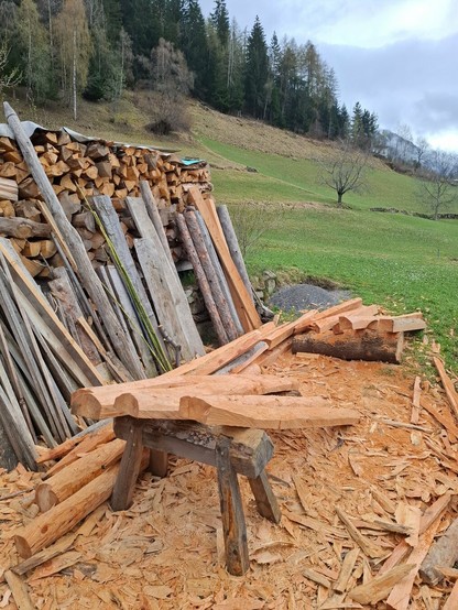 A rural outdoor woodworking area set against a wood backdrop. In the foreground, freshly stripped logs—mostly larchwooden fence posts with their sapwood copped—are scattered around a sturdy wooden sawhorse. Wood shavings and bark chips densely cover the ground. On the left, a neat stack of similarly prepared timber leans against a structure. A rustic wooden fence runs along the right side, marking the edge of a grassy field. In the background, gently sloping meadows lead to forested hills and cloud-draped mountains. The overcast sky casts a soft, diffuse light, giving the scene a calm and earthy feel.