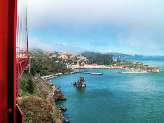 Golden Gate Bridge and Ft Baker

Captured from the north tower of the Golden Gate Bridge. 
The Golden Gate Bridge frames the left side of the image. Directly below the bridge you can see a service road that leads out to a lighthouse below the bridge.
Next to the service road is the beautiful blue waters of the San Francisco Bay. The bay runs up along the right side of the image. 
In the center of the image is Horseshoe Bay and Ft. Baker. You can see the Marin Headlands in the background. The sky is almost covered in low clouds and fog except for a somewhat circular opening above Ft Baker showing a beautiful blue sky.