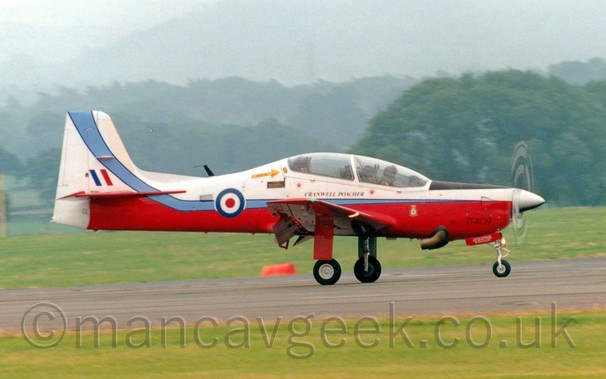 Side view of a white and red, single propellor-engined military training aircraft moving from left to right along a runway.
Flaps are extended behind the wings at an extreme angle, suggesting it has just landed.
The plane has white upper surfaces, with a red belly and wings, and a pale blue stripe running along the body which sweeps up into the white tail.
The black serial "ZF409" is in a non-standard position on the lower front of the engine cowling, with black "Cranwell Poacher" titles under the heavily glazed bubble canopy.
There is a red, white, and blue roundel on the rear fuselage, and a blue, white, and red fin flash on the tail.
Grass lines the sides of the runway in the foreground, with more grass leading off to trees in the distance, under hazy grey skies.