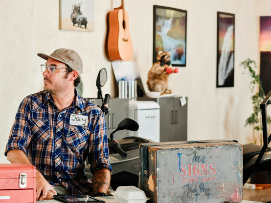 Man in room with artworks and a guitar hanging on the walls. He's looking to one side and sat in front of a table/desk that has a sign kit box on it that is lettered with "My signs don't cost, they pay".