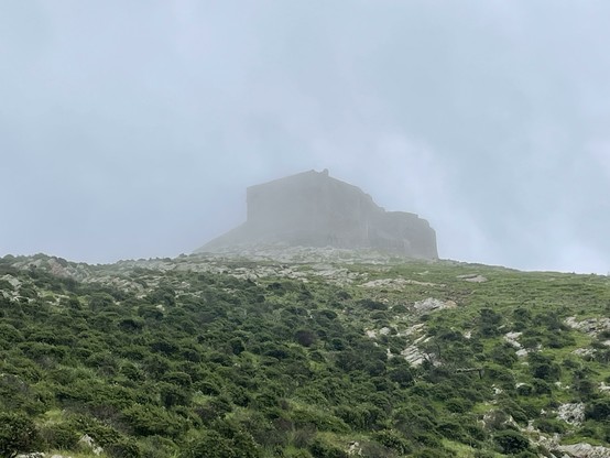 hill with green shrubs in the foreground, castle shrouded in clouds in the background