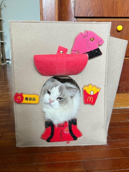 A long haired white and grey cat looking out of a felt looking bag with McDonald’s patches on it
