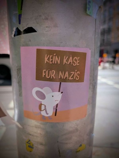 Ein Aufkleber einer weißen Maus auf rosa Hintergrund. Die Maus hält ein Schild hoch, auf dem "KEIN KÄSE FÜR NAZIS" steht.