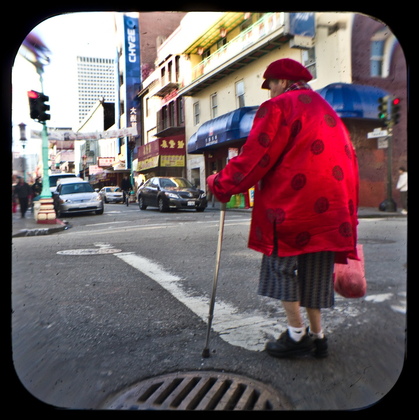An intersection in Chinatown, with a woman in a bright red coat, holding a cane, crossing the street. The scene in the distance includes restaurants, a Chase bank, streetlights, and banners spanning the street.