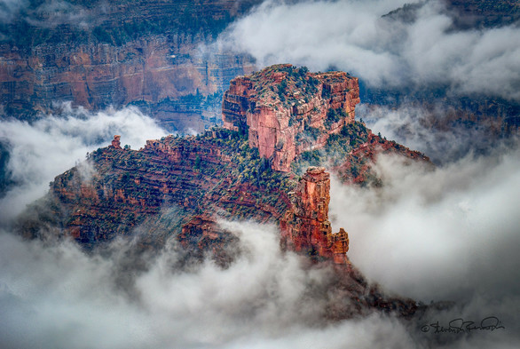 A distant view overlooking a red rock mesa and foreground rocky spire, surrounded in clearing clouds, viewed from the North Rim of the Grand Canyon