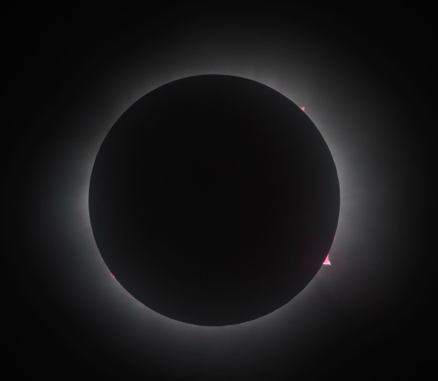 A picture of the sun being completely blocked by the moon as it passes between the earth and the sun. Around the edge of the moon, which only appears as a completely black disc, a corona of white light forms a halo. At a few places you can see a small red orange flare caused by the sun ejecting material from its surface. 