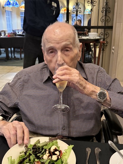 A 100-year-old man (though you wouldn't know from the photo(, sitting at a dining table. There's a yummy-looking salad in front of him and he's drinking a glass of champagne (or sparkling wine, more likely). 