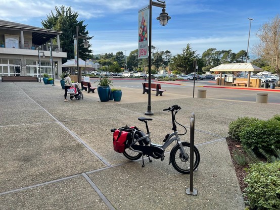 A blue Tern NBD with red panniers is parked momentarily alongside the only bicycle parking rack near the MST shuttle bus stop at Del Monte Center.  MST shelters and benches are visible in background, along with a woman pushing a baby stroller. 