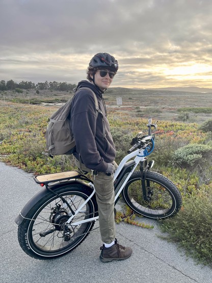 A young man stands astride his e-bike, a white-frame Himiway with a black rear afender and a wooden rack above the rear tire. He is dressed in beige pants, brown hoodie and boots, has a beige backpack on his back, and is also wearing sunglasses and a black bicycle helmet. He is on a paved multiuse trail, and behind him are the plants and trees of the Fort Ord Dunes. The sun is beginning to set. 