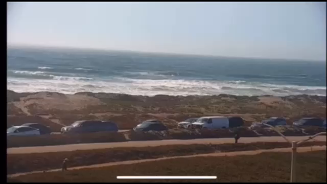 A video of the Great Highway in the afternoon, Ocean Beach and the Pacific Ocean are visible in the background. A group of people on bikes rolls slowly up the northbound side, with cars following behind. Some traffic remnants can be seen on the southbound side.