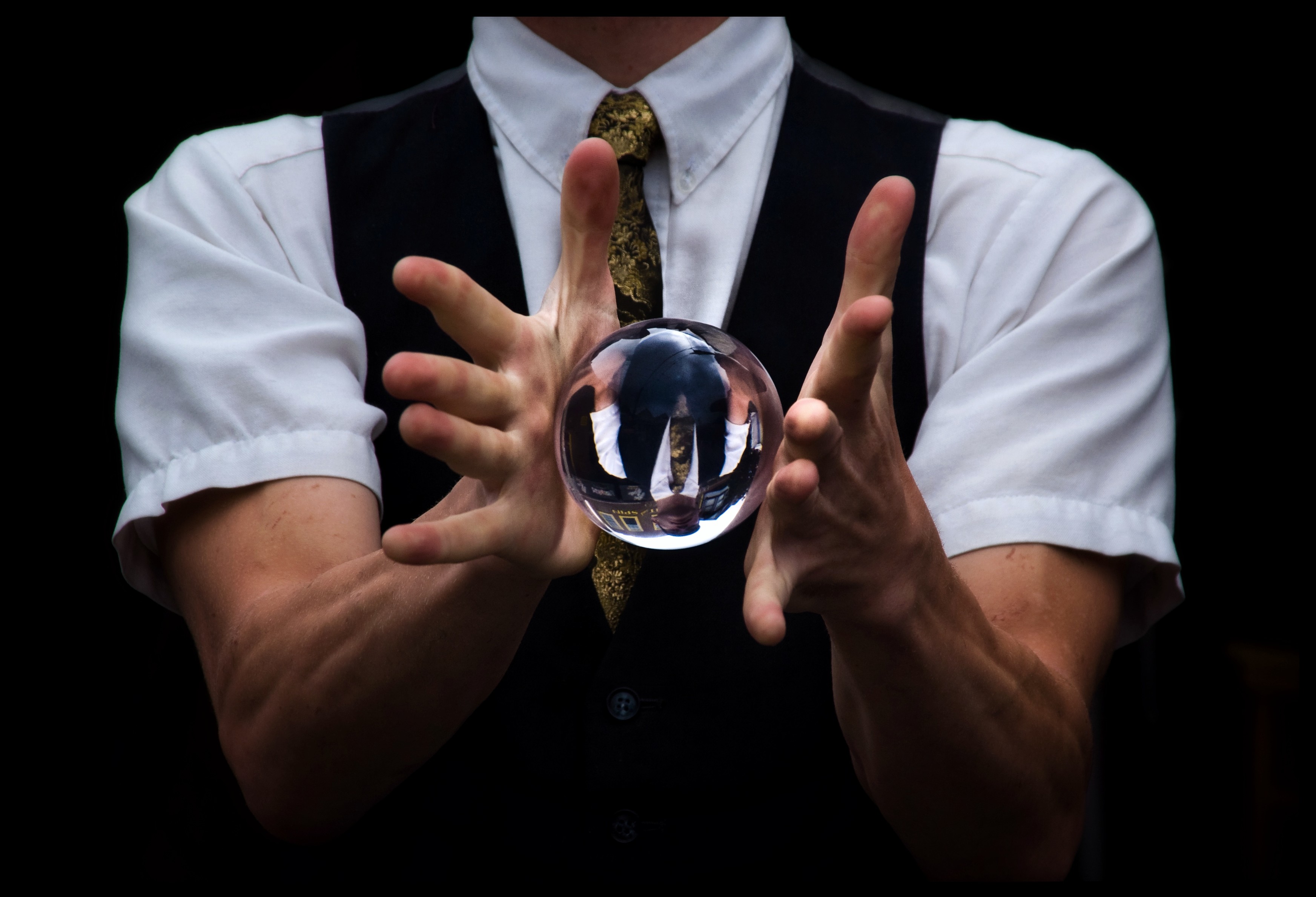 A street performer in a white shirt, dark vest, and golden tie holds a crystal ball in front of him between his two hands. His fingers are stretched out in the ball balances momentarily between his palms. A street performer in a white shirt, dark vest, and golden tie holds a crystal ball in front of him between his two hands. His fingers are stretched out in the ball balances momentarily between his palms.