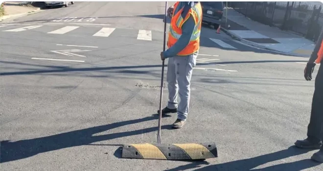 image of a person in a high-vis vest holding a long metal pole that reaches under a speed bump in an intersection, as if to pry it up