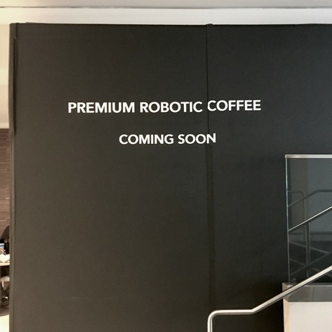 Photograph, in desaturated black and brown tones, of what appears to be temporary drywall paneling enclosing an area of a mall or other interior space that is undergoing construction or renovation. The paneling is painted black and is stenciled with white stark, sans serif lettering that reads “PREMIUM ROBOTIC COFFEE COMING SOON.”