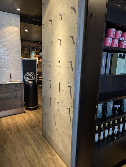 Photo of a wall in a coffee shop. On the left is a white tiled wall above a counter with a water dispenser.  In the center is a concrete wall  with a 4 wide by 5 high grid of bolts starting about 1 m above the floor, bolts spaced maybe 30 cm apart.  Each bolt has a nut threaded onto it so that each bolt has  about 5 - 10 mm sticking out.  On the right are some dark shelves displaying merchandise