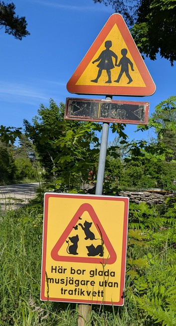 A photo of a rural road in Gotland, Sweden, with a segment of grey road to the left, green trees, grasses, and ferns under a blue sky. In the middle is a sign post with 2 signs. The top is a yellow triangle with a red border, and in the middle are the black silhouettes of a girl and a smaller boy walking hand in hand.

Beneath that is a square yellow sign with a red outline, and in the middle, surrounded by a red triangle , are the silhouettes of 3 kittens playing with butterflies.  Below are t…