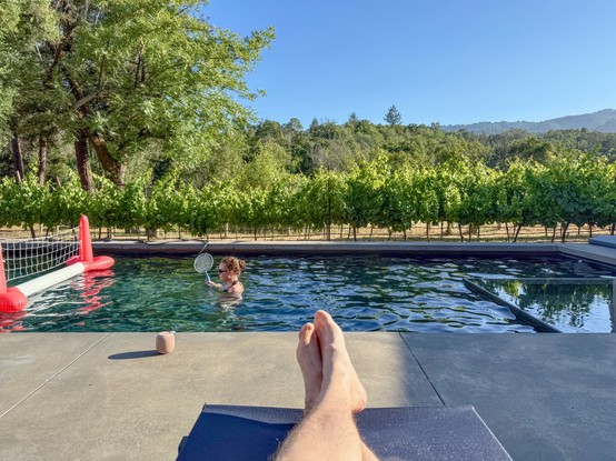 Photograph of a swimming pool and deck, emphasizing the camera’s first-person point of view. In the foreground, the photographer’s legs and feet are visible stretched on a lounge chair. A teenage girl is standing, mostly submerged, in the pool, holding a racket and playing a game like badminton. Beyond the pool, a small vineyard, large mature olive trees, and distant hills are visible in the background, all bathed in sunlight.