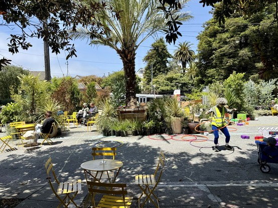 Photograph of an urban street closed to traffic and outfitted with hundreds of potted plants and trees, yellow cafe tables and chairs, and physical playthings including #HulaHoop rings and giant #Tetris blocks. Several people are seated or hula hooping in clear and dappled sunlight, while a light rail train is partially visible in the background behind the parklet.