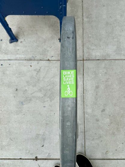 Photograph, looking straight down at a concrete sidewalk, of a bright green sticker affixed to the top of a circular #bicycle rack. The sticker reads, “Bike lanes save lives 🚲.”