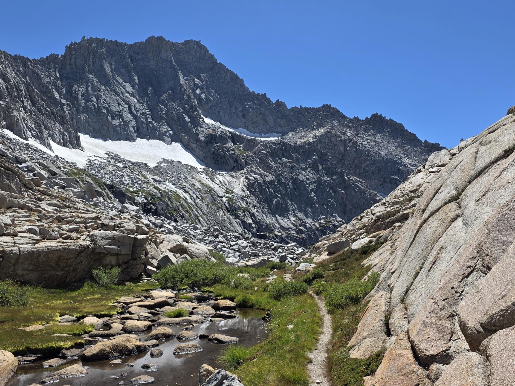 A trail leading around a corner to a lake with a tall cliff face of a mountain rising behind