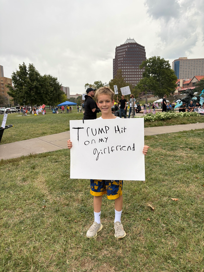 A young white boy, maybe 10’or 11 years old standing on grass holding a white paper sign with the words “Trump hit on my girlfriend” 
