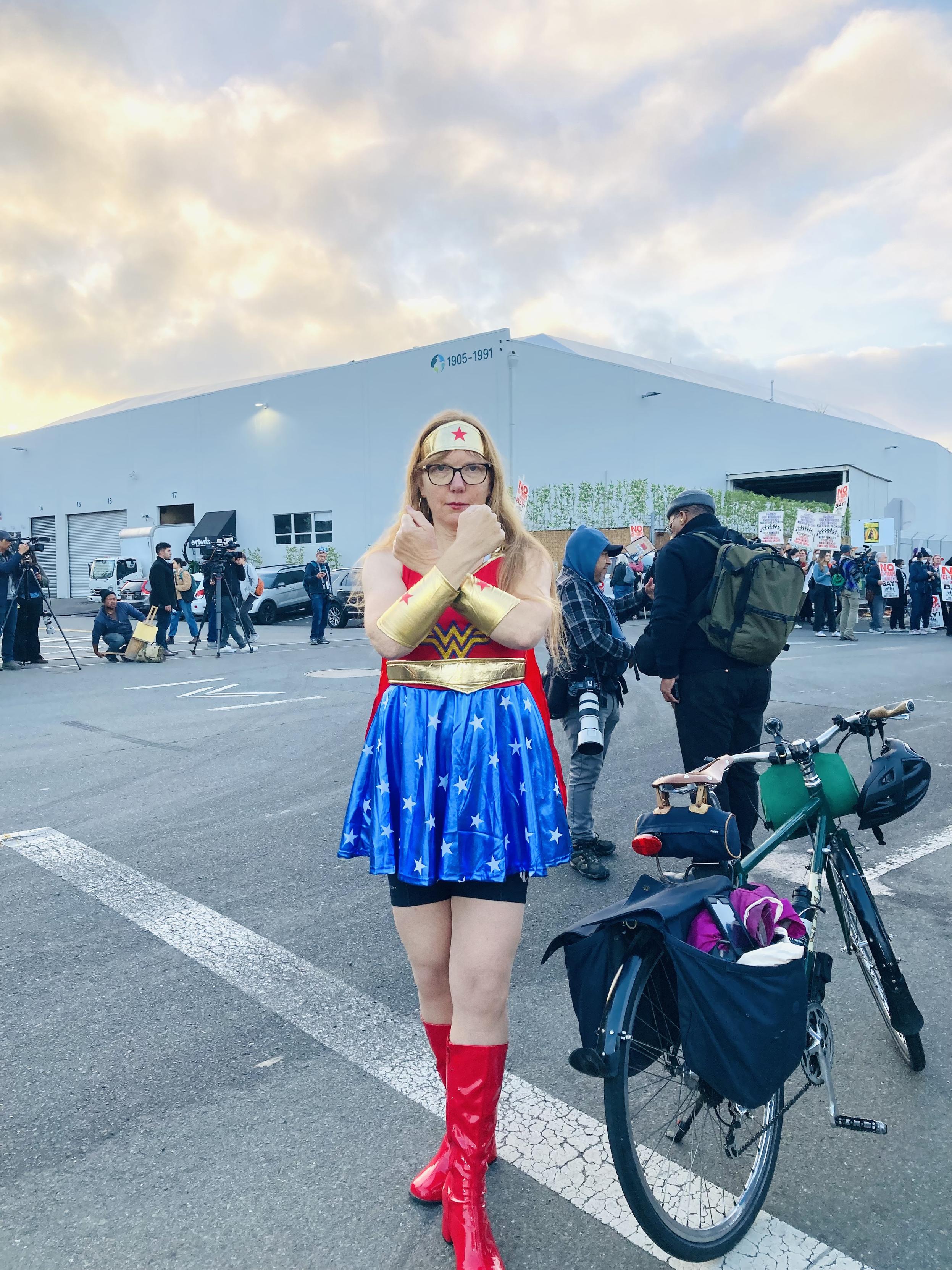 A photo of a woman dressed as Wonder Woman with her arms crossing her chest, standing next to her bicycle at a protest.