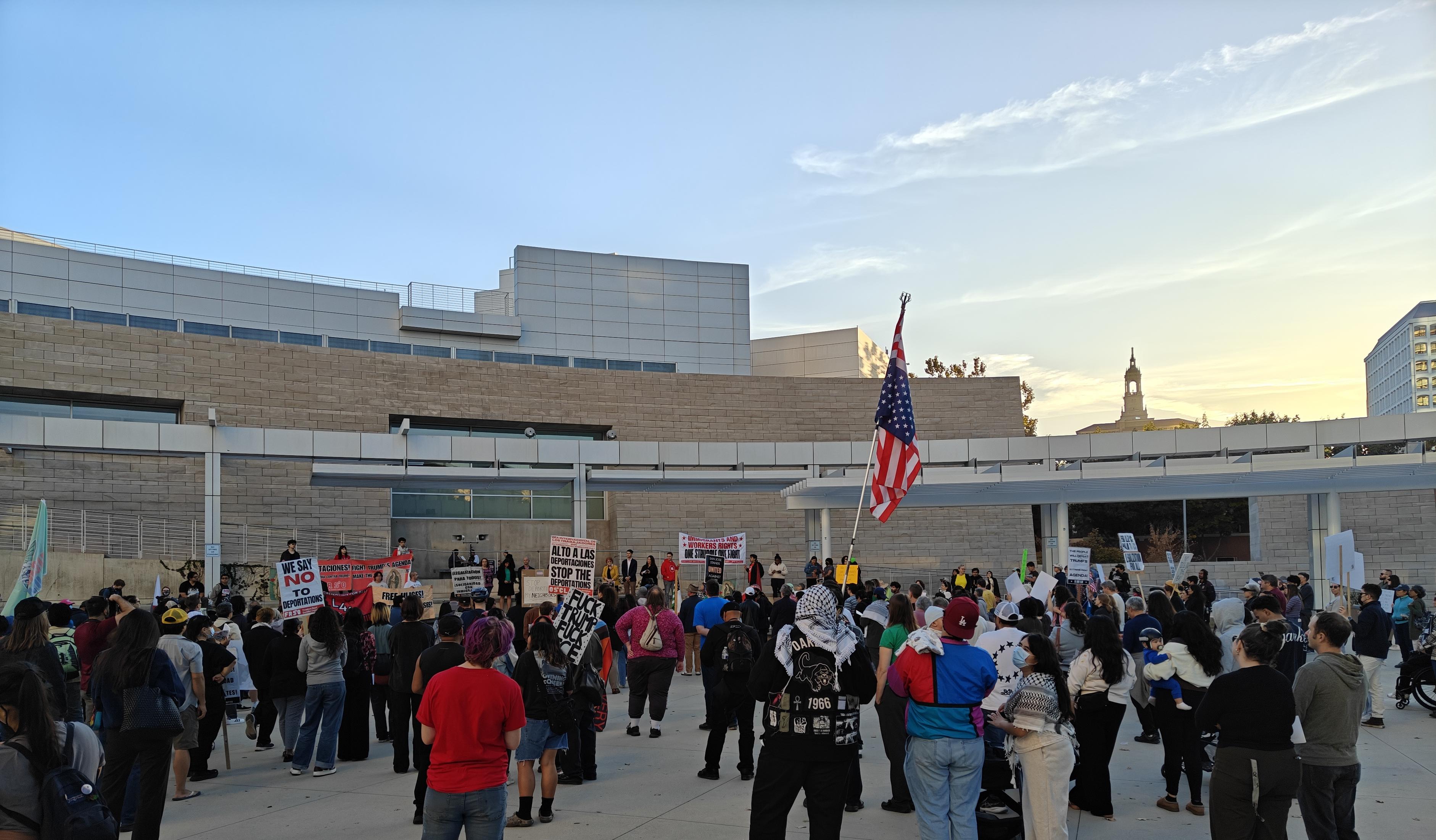 A large, varied group of people at a protest, seen from the back, many holding signs and one with a U.S. flag flying upside down.