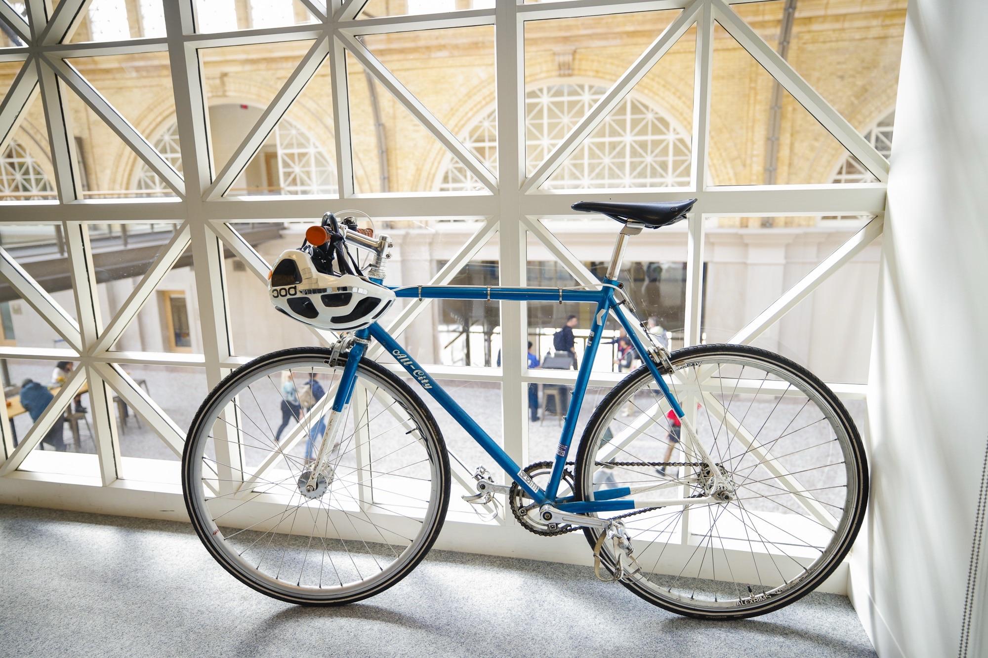 A blue bicycle is leaning against a window with a geometric frame. A helmet is attached to the handlebars. Through the window, people can be seen walking in a spacious area with architectural features.