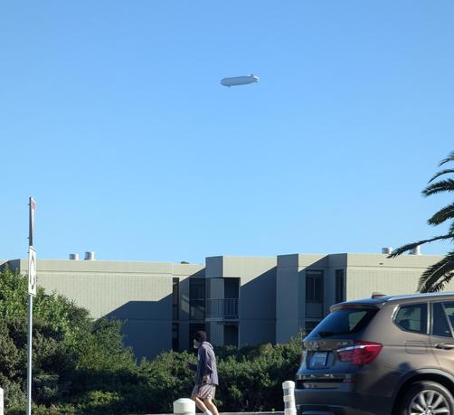 A photo of a blue sky above a concrete building, with greenery, a pedestrian, a car, a road sign and some bollard tops in the foreground. Centered toward the top is a large dirigible ( or blimp?)