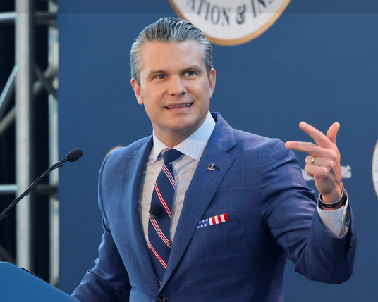 A photo of Pete Hegseth at a podium wearing a blue suit, a darker blue tie with widely spaced bars of red and white stripes, and a folded pocket handkerchief in the breast pocket of the suit.  The handkerchief shows part of a flag - with a blue rectangle showing 2 rows of stars on the left,  and 3 white and 3 red stripes to the right.