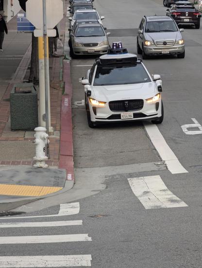 Photograph of a Waymo, blinkers flashing, pulled most of the way in to the red painted curb of a bus stop, which is clearly identified by the yellow band (and blue sign, not visible in photo).  You can see the letter P on the pavement to the right of the Waymo (left from photos view).  The car is stopped over the "BUS STO" painted in the stop.