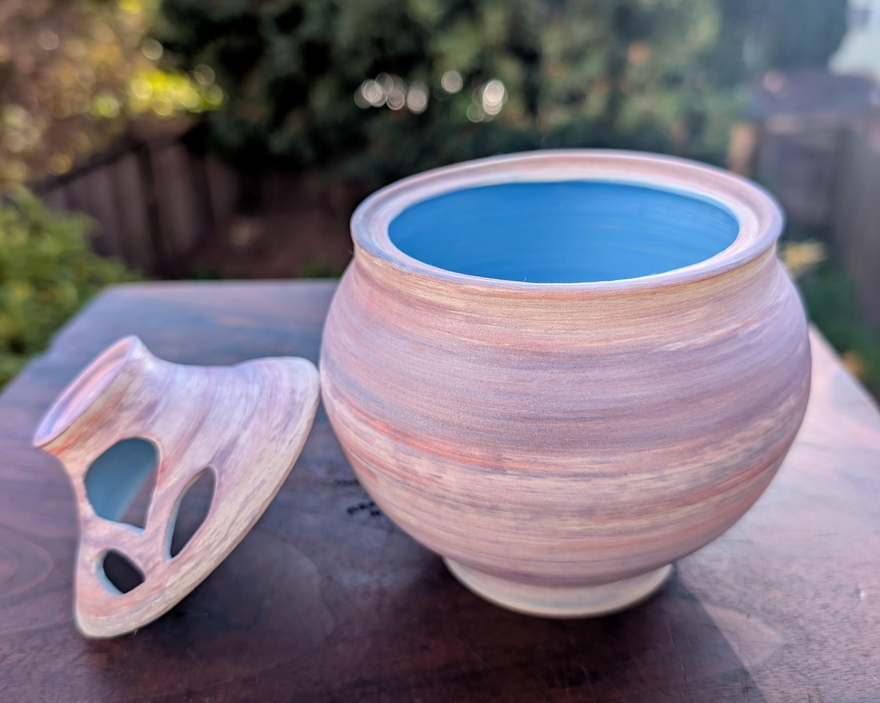 A wheel-thrown bulb-shaped pot, with the lid resting against the pot. The lid has holes in it to let the garlic breathe. The exterior of the pot is underglazed in streaks of white, purple, and pink, reminiscent of purple garlic. The interior is the pot is a bright azure. CC BY-SA 4.0 Kate Zimmerman.