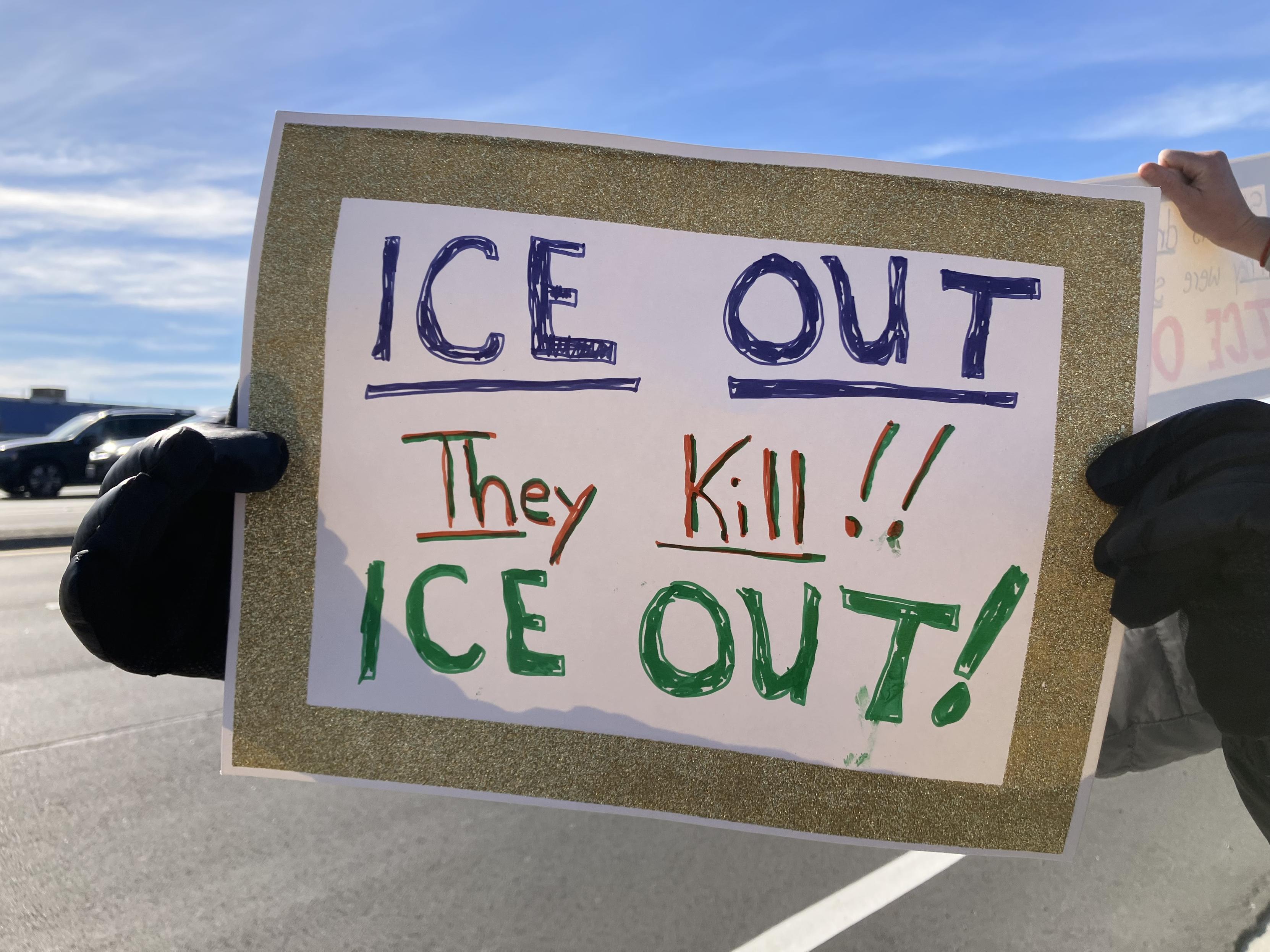 Someone holding a sign, about 8.5 inches by 11 inches, which says:
ICE Out
They Kill!
ICE Out!

The sign has a border of gold glitter. 
