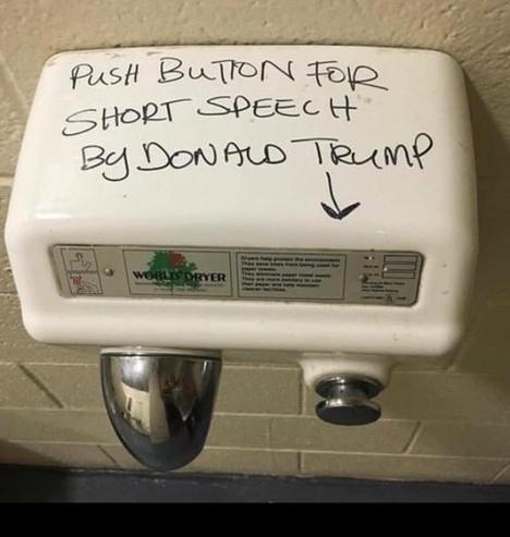 A photo of a hot air hand dryer mounted on tile wall, viewed from above.  There's a chrome air vent on the left, and a large push button on the right.  In black, on the dryer's top, someone has drawn an arrow pointing to the button, and behind that, in capital letters, has printed "PUSH BUTTON FOR SHORT SPEECH BY DONALD TRUMP"